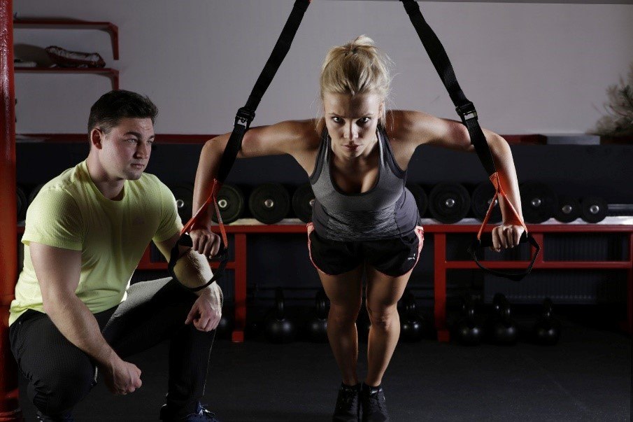 Female is exercising in gym with male trainer watching